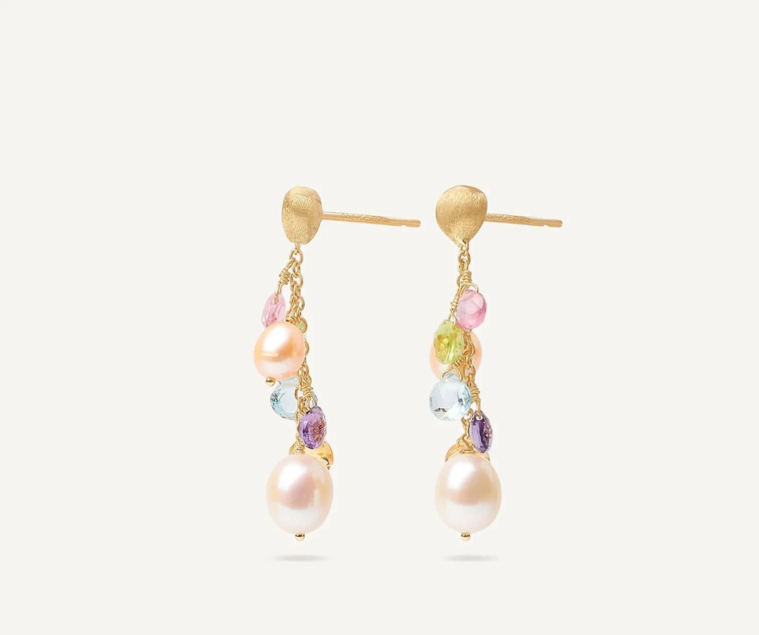A pair of gold dangle earrings featuring large white freshwater pearls, small multicolored gemstones, and textured gold posts, shown against a light off-white background. angled