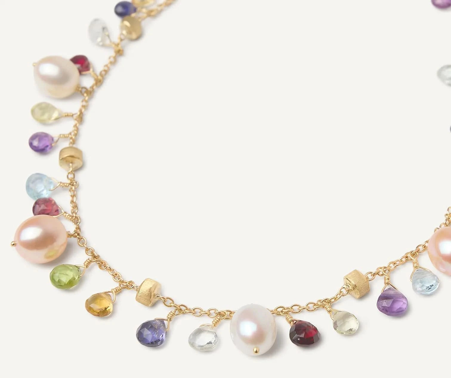 A delicate gold chain necklace adorned with small multicolored gemstones and white freshwater pearls, arranged in a circular shape on a light cream background. close up