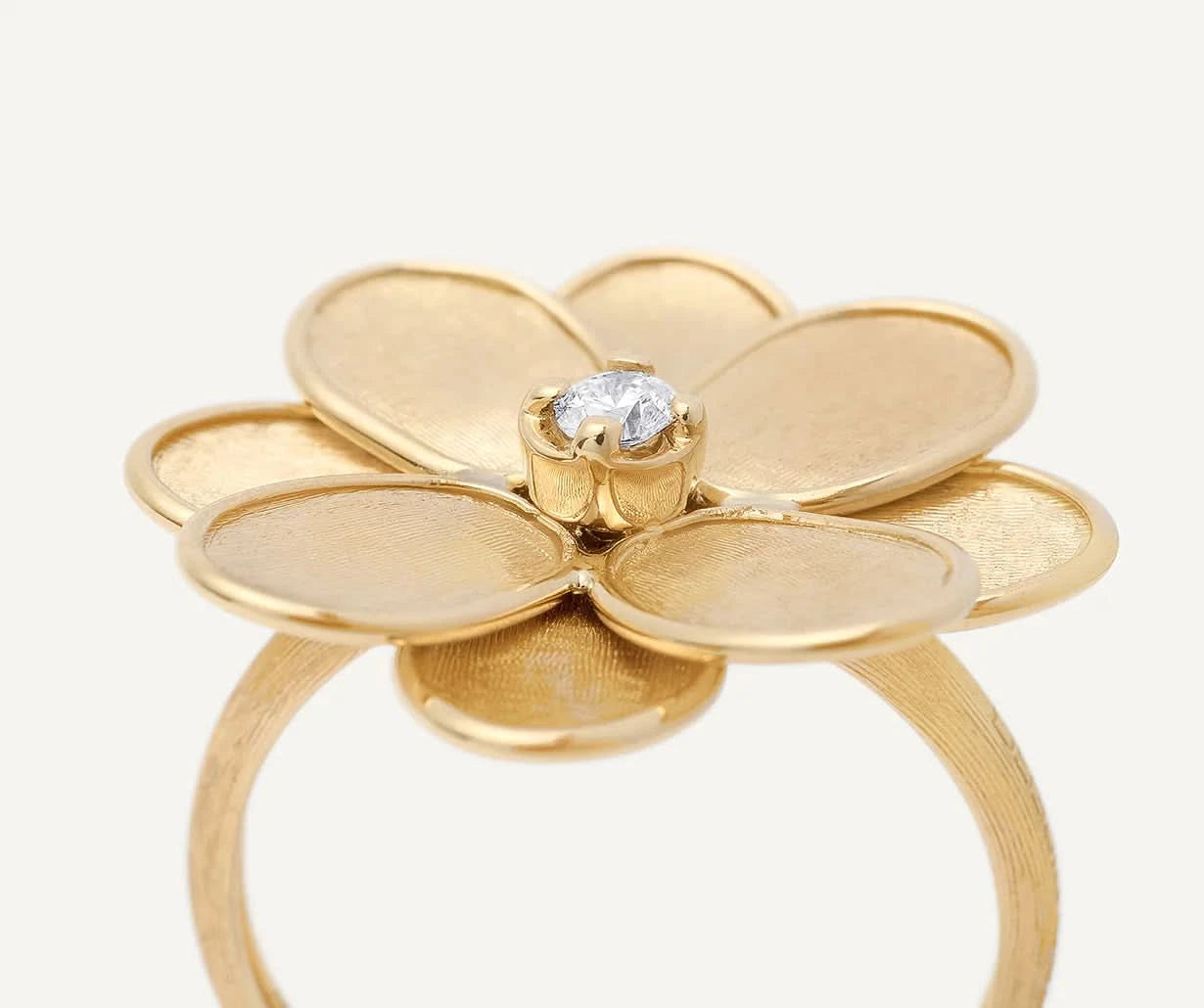 A gold flower ring with five textured petals and a central clear gemstone, shown on a light off-white background. Close up view.