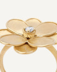 A gold flower ring with five textured petals and a central clear gemstone, shown on a light off-white background. Close up view.