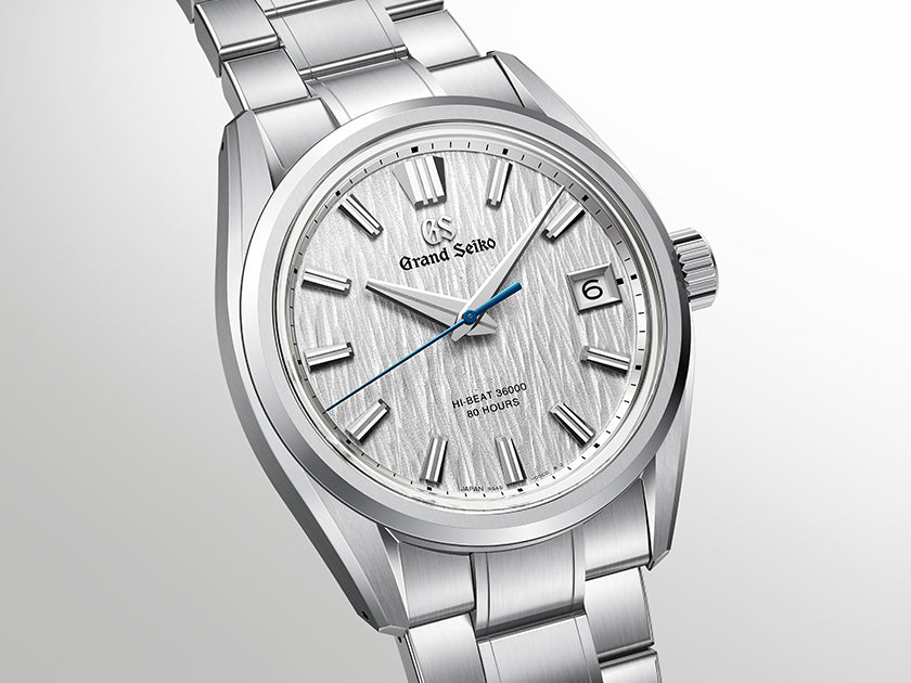 A Grand Seiko stainless steel wristwatch with a textured silver dial, silver-tone metal bracelet, and a distinct blue second hand, shown against a light gray gradient background.