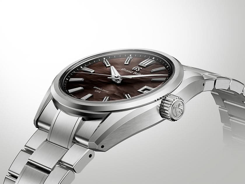 A close-up studio shot of a wristwatch with a silver-toned metal bracelet and case, featuring a dark brown dial with silver baton markers and a textured crown.