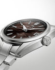 A close-up studio shot of a wristwatch with a silver-toned metal bracelet and case, featuring a dark brown dial with silver baton markers and a textured crown.