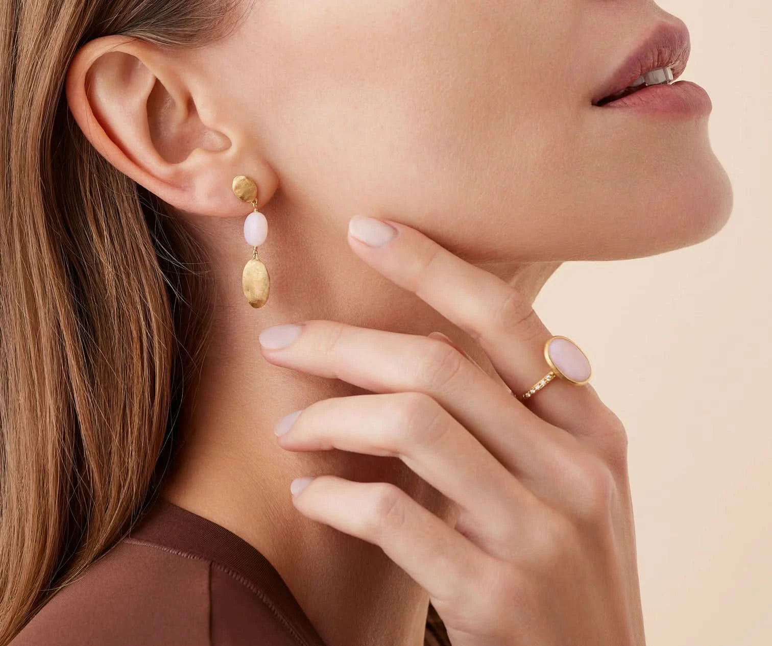 A close-up lifestyle shot of a model wearing a gold drop earring with pale pink oval beads and a gold ring with a pale pink oval stone, against a soft beige background.