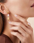 A close-up lifestyle shot of a model wearing a gold drop earring with pale pink oval beads and a gold ring with a pale pink oval stone, against a soft beige background.