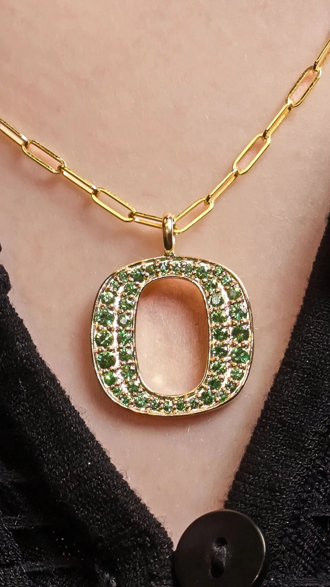 Gold chain necklace with a large open letter 'O' pendant encrusted with green gemstones, shown worn on a person's neck.