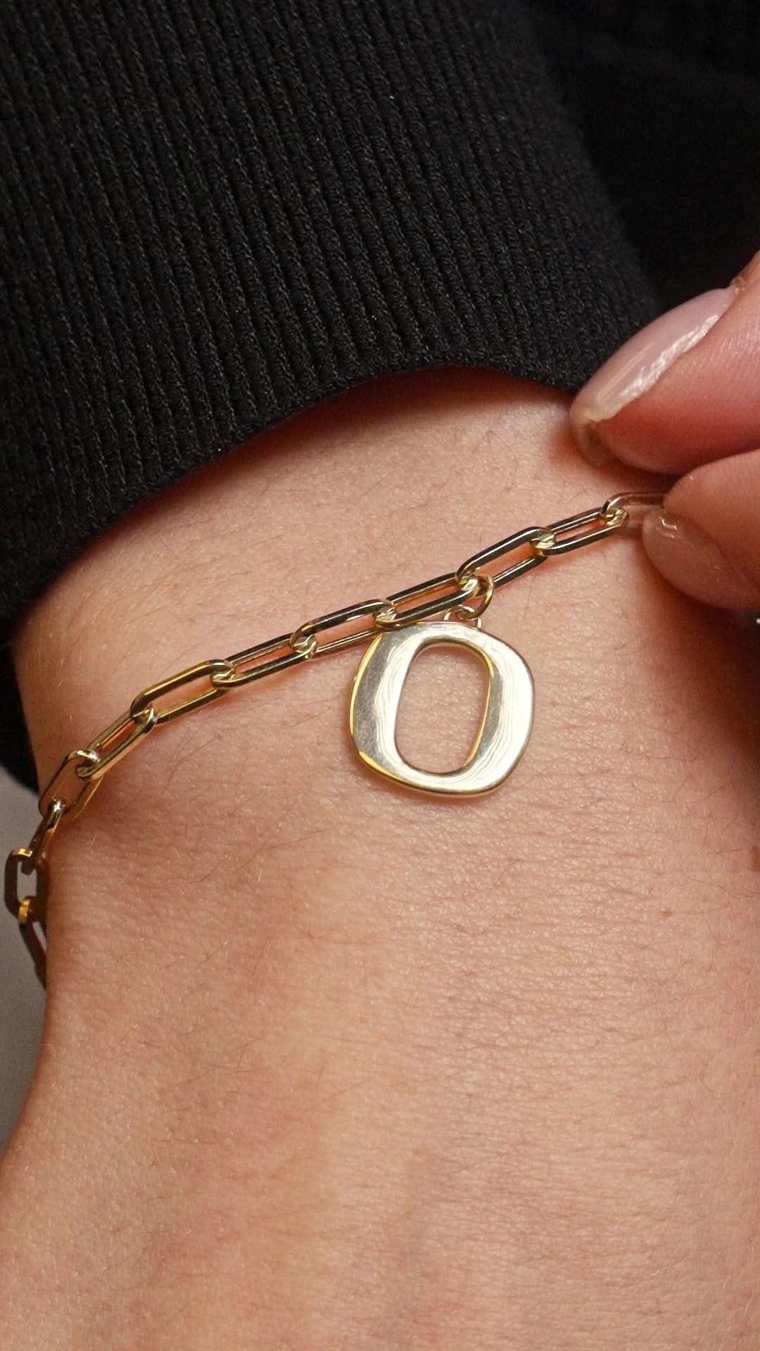 A close-up of a gold chain bracelet with a polished oval letter 'O' pendant, worn on a person's wrist with a black ribbed sleeve visible.