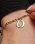 A close-up of a gold chain bracelet with a polished oval letter 'O' pendant, worn on a person's wrist with a black ribbed sleeve visible.